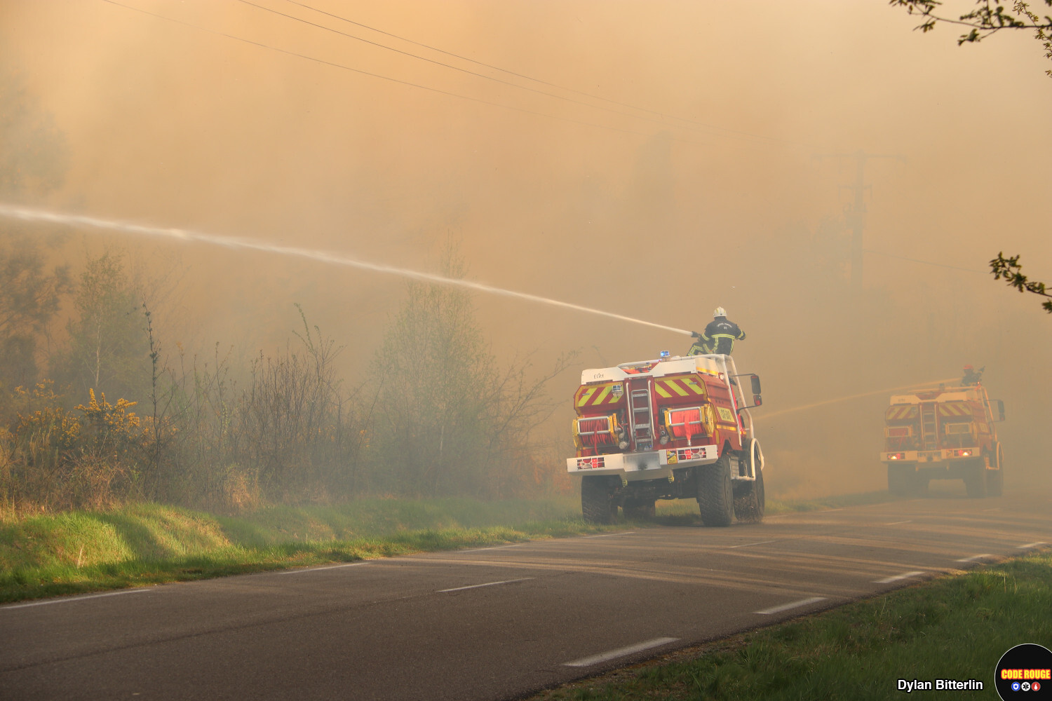 Deux véhicules de sapeurs-pompiers combattent les flammes depuis le bord de la route, au milieu de la forêt