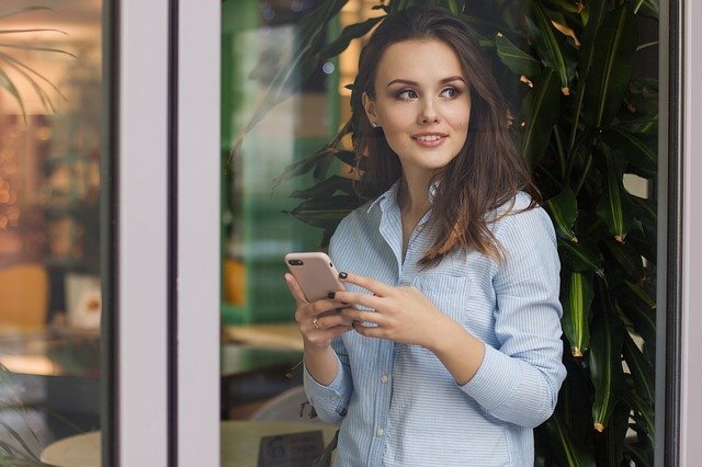 Femme portant une chemise bleue regardant par la fenêtre avec un téléphone en mains