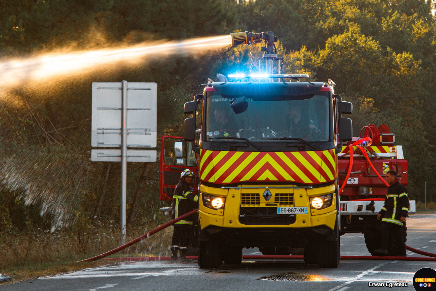 Des véhicules de sapeurs-pompiers luttent contre les flammes depuis le bord de la route ; deux sapeurs-pompiers se tiennent debout à proximité sur la route