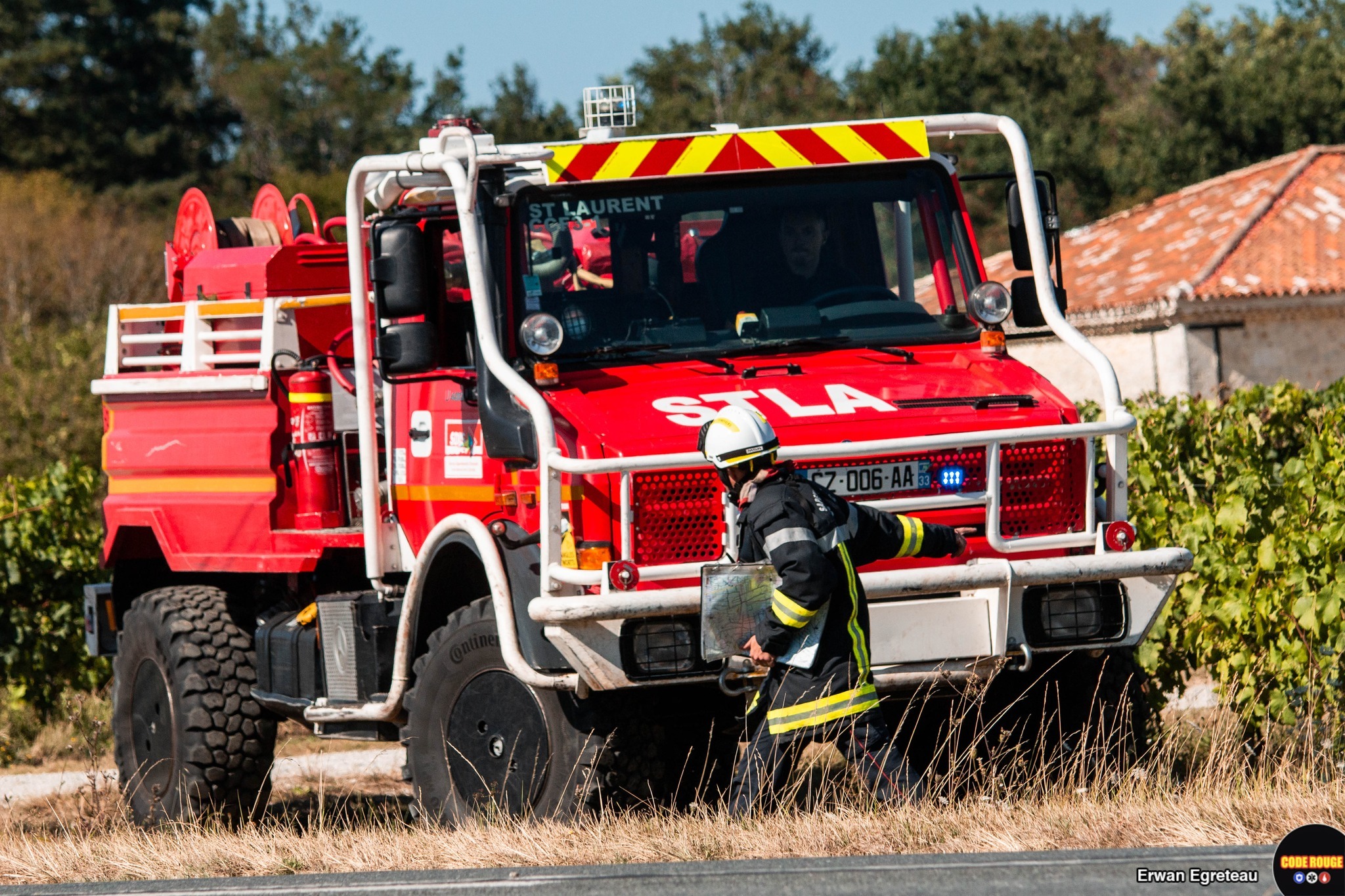 Un sapeur-pompier de profil en uniforme tenant une carte dans la main gauche indique une direction vers la droite à un véhicule de sapeurs-pompiers en face de lui