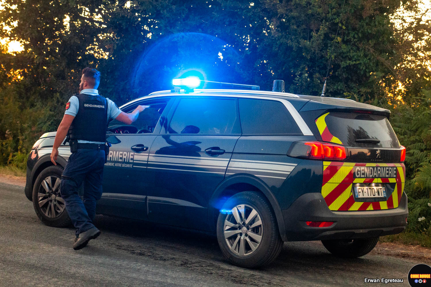 Un gendarme de dos, fait un signe à son collègue dans une voiture de gendarmerie à côté de lui