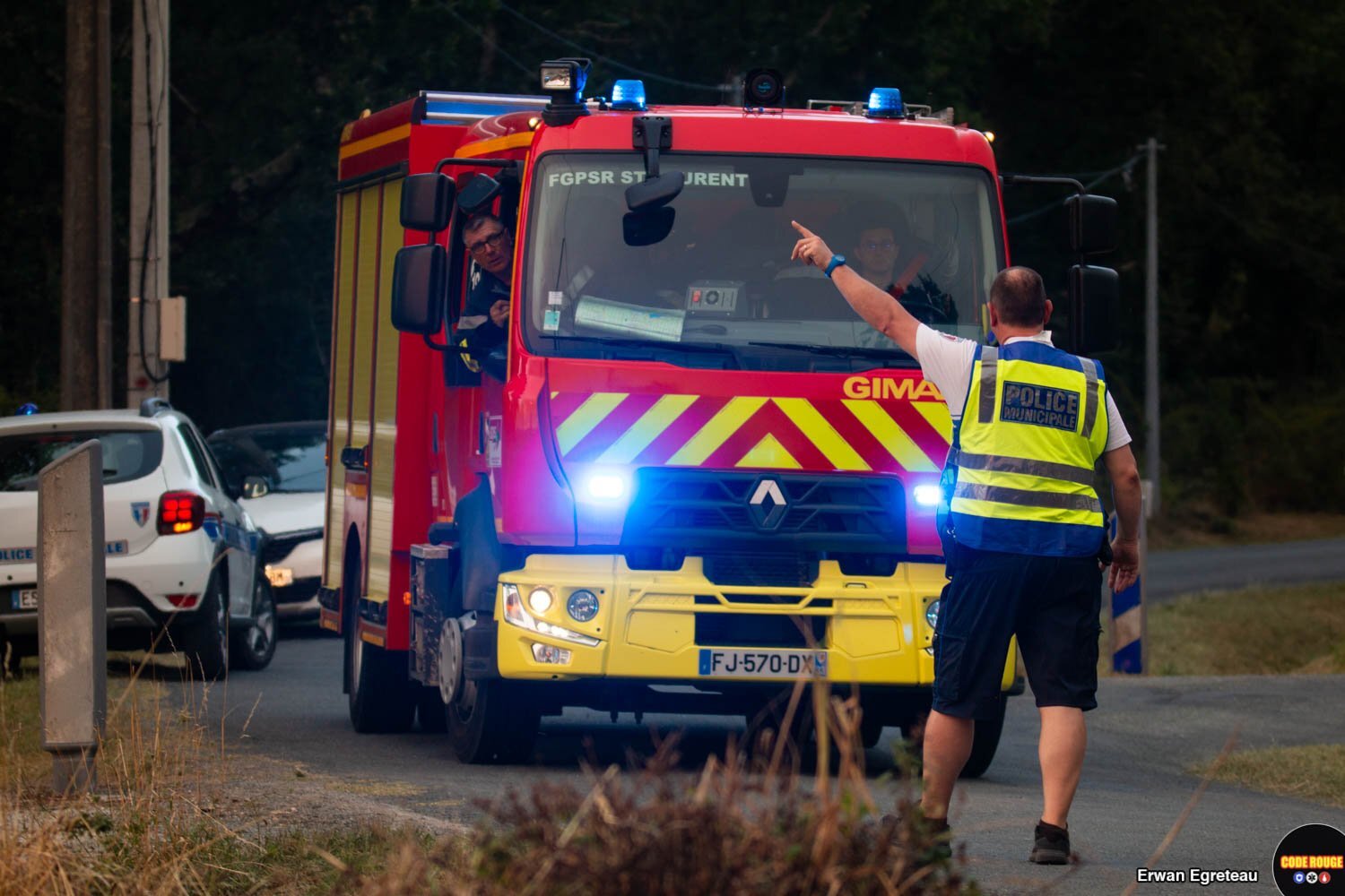 Un agent de la police municipale, de dos, indique une directio vers la gauche à deux sapeurs-pompiers qui arrivent vers lui dans un camion