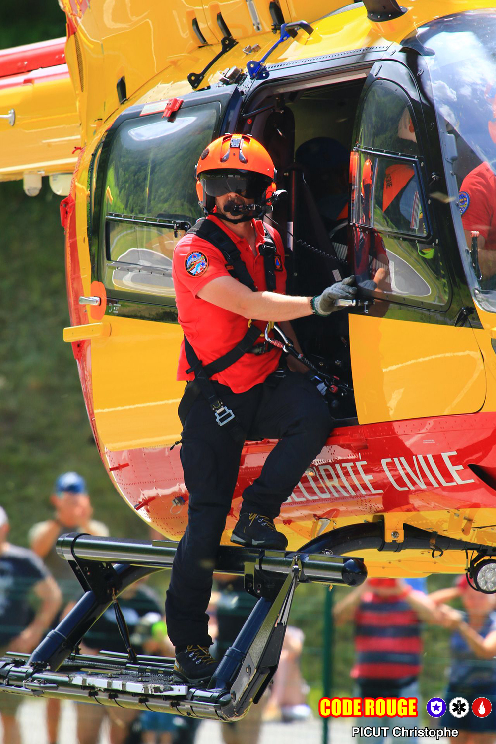 Un hélicoptère de la Sécurité Civile proche du sol lors d'une démonstration ; un agent se tient débout sur les patins de l'hélicoptère