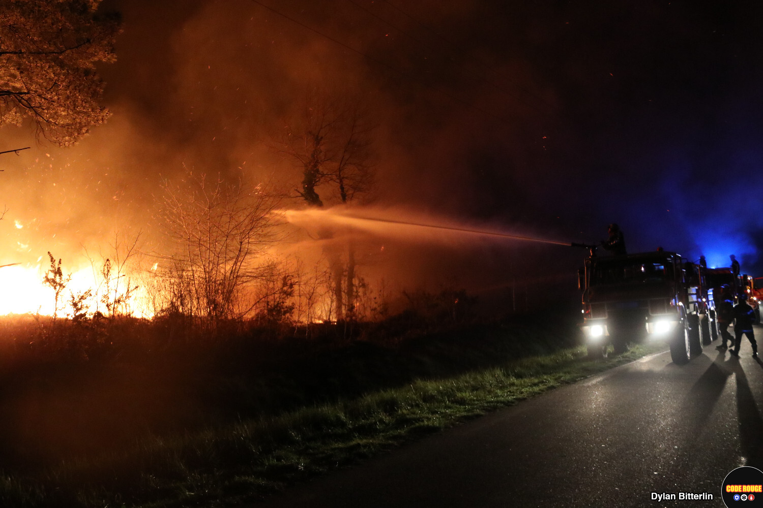 Plusieurs sapeurs-pompiers luttent contre les flammes depuis la route, en pleine nuit
